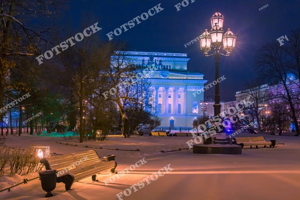 На фотографии запечатлен Александринский театр ночью в зимнее время. Заснеженный сквер перед театром украшен изящными старинными фонарями, которые мягко освещают белоснежные лавочки и деревья. Здание театра, подсвеченное холодными тонами, выделяется своей классической архитектурой. Атмосфера спокойной зимней ночи наполняет кадр гармонией и элегантностью.